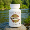 AyuMantra Haritaki Tablets bottle on a rock by a river, dietary supplement label visible.